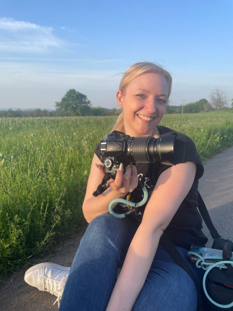 lächelnde Fotografin mit Kamera in der Hand vor einer Blumenwiese bei Sonnenschein